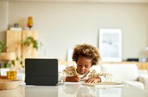 A child writing on paper with a tablet computer on the counter nearby.