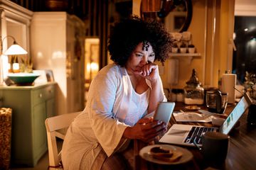 A woman sitting at a desk in front of her laptop looking at her smartphone