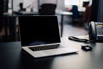 A powered-off MacBook Pro sitting on a desk in an office environment with an office phone and mouse nearby.